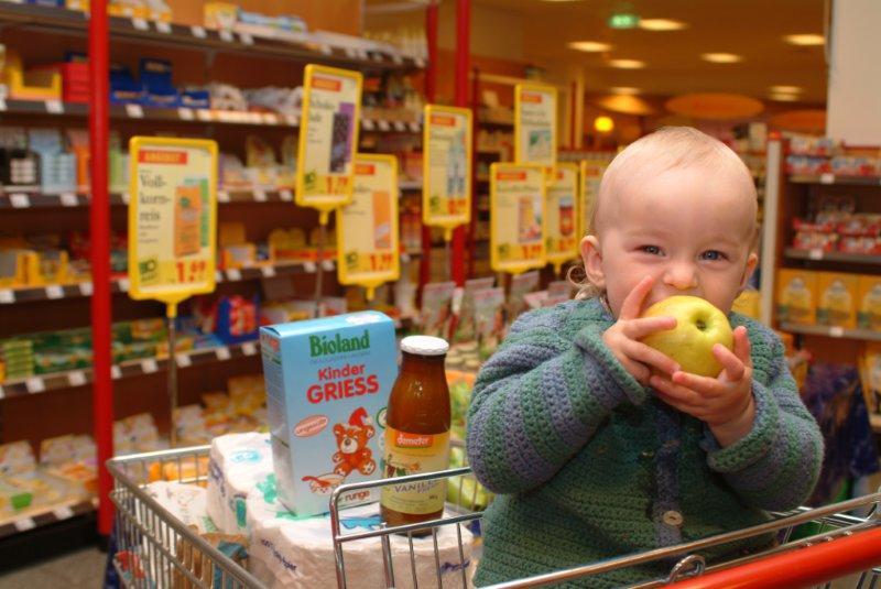 Ein Kleinkind im Einkaufswagen im Supermarkt beißt zufrieden in einen Apfel. Klick führt zu Großansicht im neuen Fenster.